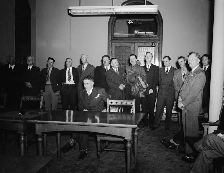 In this black and white photo, a group of about a dozen men in suits stands in a courtroom.