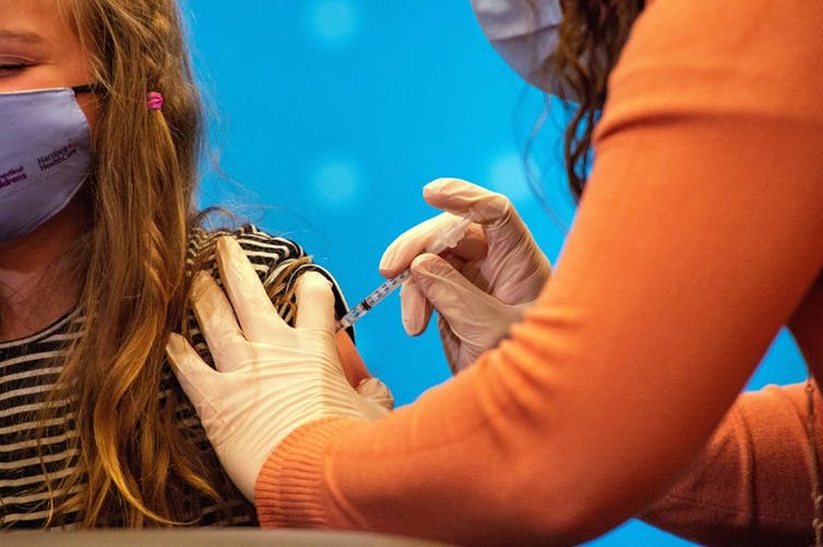 A child with long hair wearing a mask receives a Pfizer COVID-19 shot.
