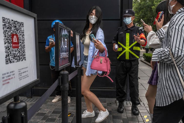 Person scans phone app used for contact tracing and has temperature checked by guard before entering a shopping area