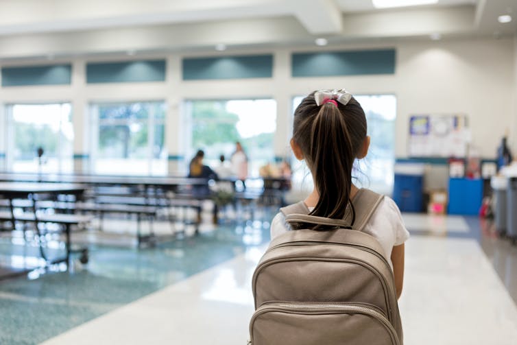 Rear view of a young girl with a backpack walking into a school cafeteria