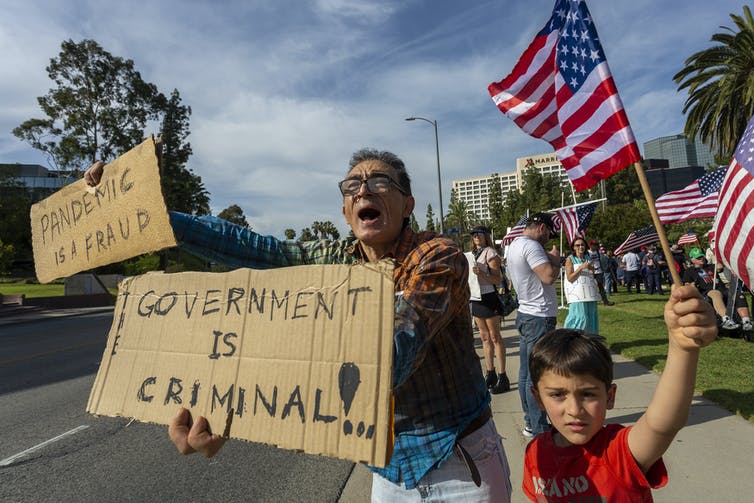 man with child holds homemade signs that read 'pandemic is a fraud' and 'government is criminal'