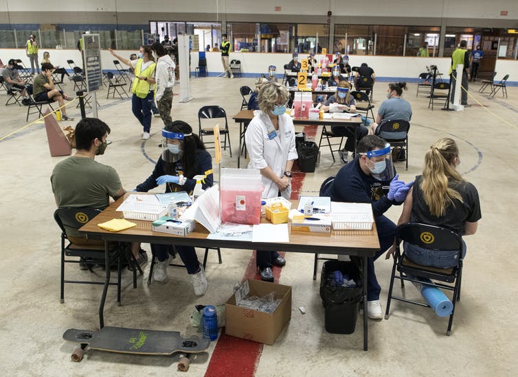 Students and medical personal at tables in a large open area.
