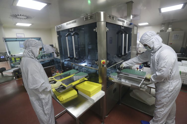 Employees in protective coveralls work on a machine filling vials with vaccine.