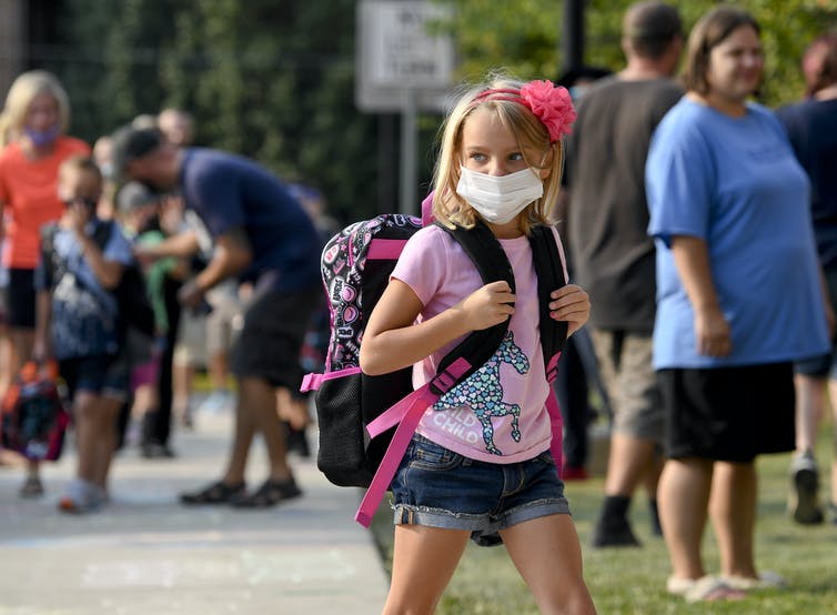 A girl wearing a mask heads to school past adults without masks.