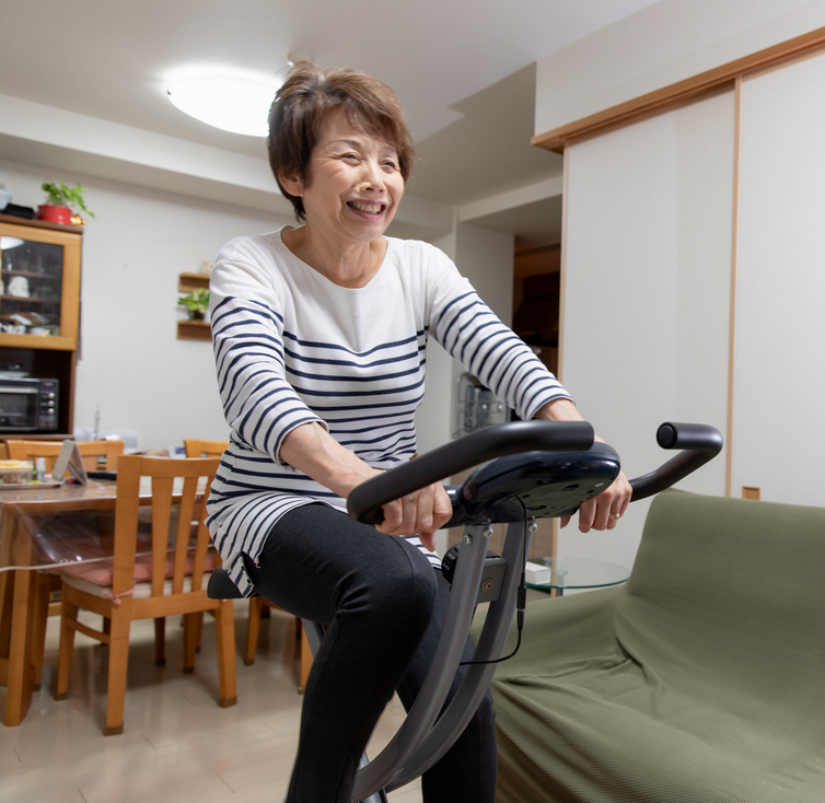 An older woman pedaling on an exercise bicycle in her living/dining room.