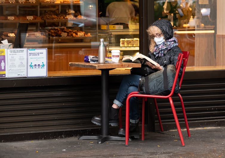 A woman in face mask and jacket sits outside a bakery.