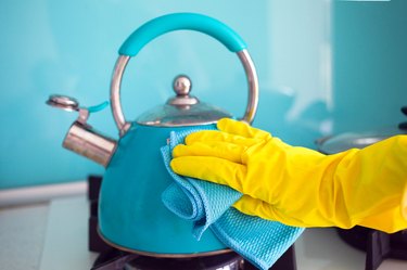 Female hand in a rubber yellow glove wipes a blue teapot with a rag