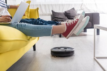 Cropped view of kid with laptop near mother with raised legs on sofa and robotic vacuum cleaner on floor in living room