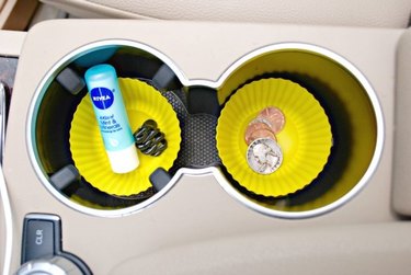 Overhead photo of two bright-yellow cupcake papers in a car's console cup holders, one containing moisturizer and one holding loose change.