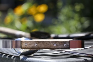 Grill tongs resting on the side table of a gas barbecue, with blurred grill brush and planter of flowers in the background.