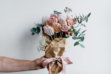 A male hand holding a bouquet of sugar cookies cut and iced to resemble flowers, wrapped in brown paper with stems of real plants to complete the bouquet.