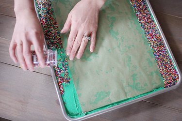 Pouring sprinkles onto sides of sheet cake while parchment paper protects center of cake