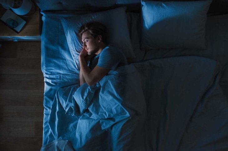 Top View of Handsome Young Man Sleeping Cozily on a Bed in His Bedroom at Night. Blue Nightly Colors with Cold Weak Lamppost Light Shining Through the Window.